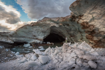A man stands in the snow in front of entrance to the snow cave of the Alibek mountain glacier in Dombay with high walls of frozen snow