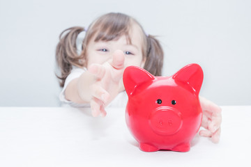 child girl pulls hands to the red piggy bank standing on the table