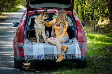 Happy Caucasian girl sitting in the trunk of a car, hugging a German shepherd dog and smiling, looking at each other. cheerful teen puppy. Friendship of man and animal, travel, camping. Autumn forest