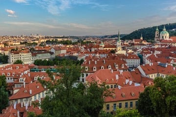 Fototapeta na wymiar Prague, Czech Republic - July 20 2019: View of red roofs, cityscape with churches and historical buildings. Blue evening sky with clouds. Green trees in foreground.