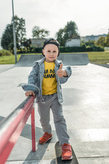 Cheerful little boy 3-5 years old, hand gesture finger to top like, great and super. Training and learning to ride skateboard. Autumn day in summer in city. Casual wear, denim pants, baseball cap.