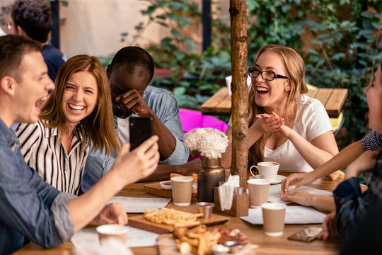Sincere laugh and showing picture on the smartphone at the casual meeting with best friends on the open air restaurant terrace