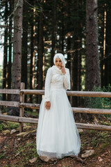 Beautiful elegant bride with hijab in lace wedding dress with long full skirt and long sleeves. Outdoors, in the woods, wooden fence in the background.