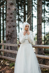 Fototapeta premium Beautiful elegant bride with hijab in lace wedding dress with long full skirt and long sleeves. Outdoors, in the woods, wooden fence in the background.