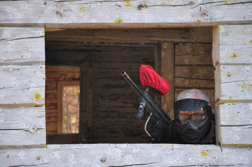 Active paintball game. A paintball player in a protective uniform and mask with a paintball marker is sitting in an ambush in an old house.