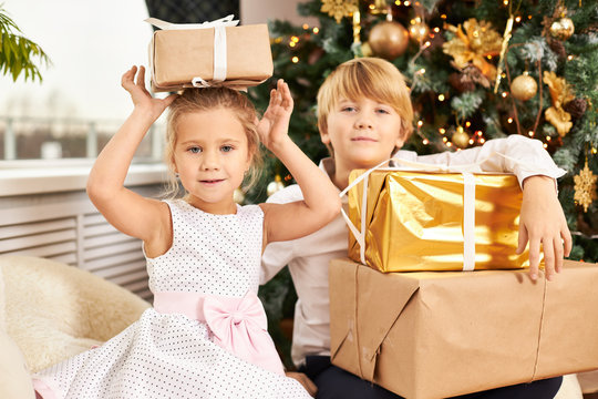 Picture of two adorable European children siblings posing at Christmas tree. Handsome teenage boy unpacking New Year’s gifts together with his cute little sister next to him with box on her head