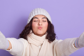 Woman making kiss gesture towards camera and spreading hands aside, keeping eyes closed, female with dark hair wearing white warm sweater, cap and scarf, posing isplated over lilac studio background.