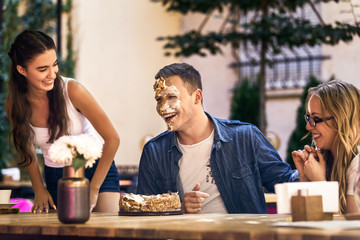 Two caucasian girls and a guy with face diry  with cake cream are laughting and sitting around the...