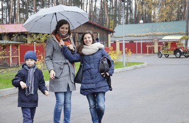 A young mother with her son and a friend happily walking in the Park. The sudden rain. Girls laugh, standing under the umbrella. Autumn, joyful walk girlfriends with a little boy.