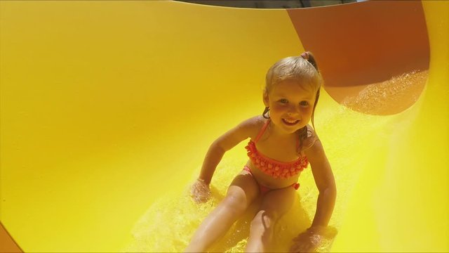 Funny little girl rides down an orange slide in a water park. Girl in a swimsuit laughs and slides into the water raising spray