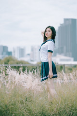 beautiful asian lady in Japanese student uniform looking at camera and standing with hand touching her hair in a beautiful grass flowers field on sunny day in summer with city background