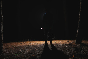 Silhouette of man with bright flashlight in forest at night - obrazy, fototapety, plakaty