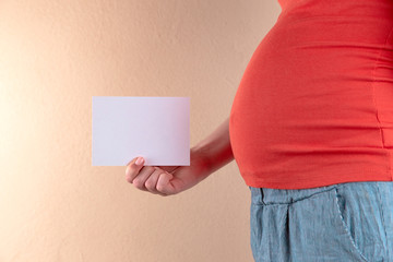 A close-up view of the belly of a pregnant woman in a red T-shirt that is holding an empty sheet of paper.