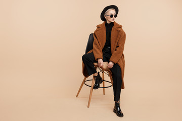 Serious young hipster woman with blonde short hair wearing a coat, hat and sunglasses posing over beige background. - obrazy, fototapety, plakaty