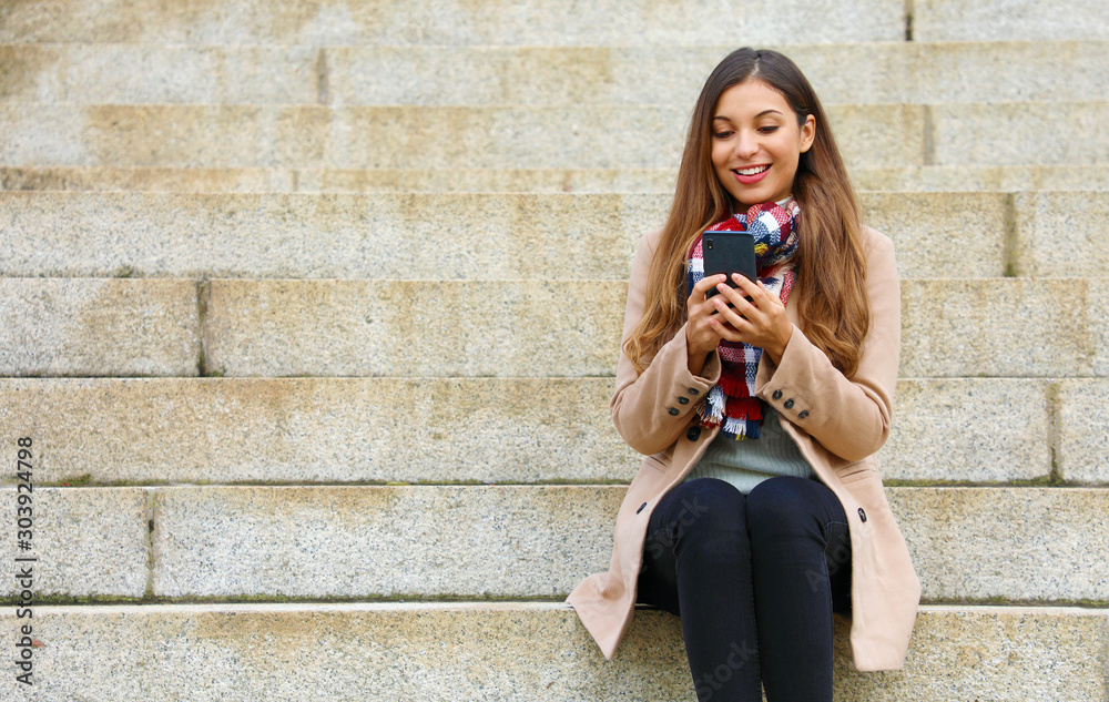 Wall mural Smiling beautiful young woman sitting on stairs using mobile phone on winter day.