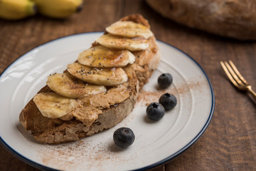 Healthy breakfast of sourdough toast with peanut butter, banana, blueberries, cinnamon, chia seeds and maple syrup. Country loaf and bananas in the background. Horizontal. Closeup