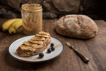 Vegan breakfast with toasted rustic bread with peanut butter, banana, blueberries, cinnamon, chia seeds and maple syrup. Country loaf, jar of peanut butter and bananas in the background. Horizontal