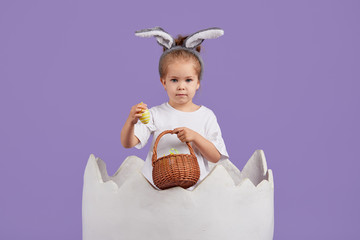 Happy child girl in costume Easter bunny rabbit with ears and a basket with eggs. Little girl in a big chicken egg on a purple background