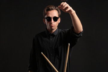 Stylish man drummer in a black shirt and sun glasses playing drums with sticks over dark background.