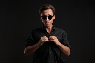 Obraz premium Confident stylish bussinesman in black eyeglasses buttoning up balck shirt isolated over black background.