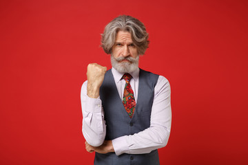 Strict elderly gray-haired mustache bearded man in classic shirt vest colorful tie isolated on red background in studio. People sincere emotions lifestyle concept. Mock up copy space. Clenching fist.