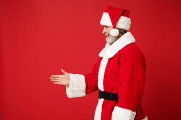 Fototapeta na wymiar Elderly gray-haired mustache bearded Santa man in Christmas hat posing isolated on red background. New Year 2020 celebration concept. Mock up copy space. Standing with outstretched hand for greeting.