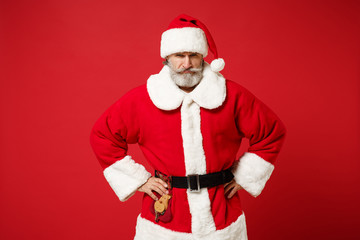 Fototapeta na wymiar Strict elderly gray-haired bearded mustache Santa man in Christmas hat posing isolated on red background. Happy New Year 2020 celebration holiday concept. Mock up copy space. Stand with arms akimbo.