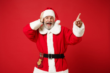 Fototapeta na wymiar Excited elderly gray-haired bearded mustache Santa man in Christmas hat posing isolated on red background. Happy New Year 2020 celebration holiday concept. Mock up copy space. Point finger aside up.
