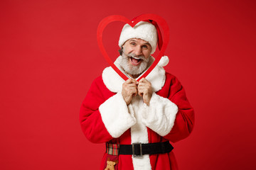 Fototapeta na wymiar Cheerful elderly gray-haired mustache bearded Santa man in Christmas hat posing isolated on red background. Happy New Year 2020 celebration holiday concept. Mock up copy space. Hold big wooden heart.