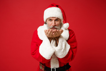 Funny elderly gray-haired mustache bearded Santa man in Christmas hat posing isolated on red background. New Year 2020 celebration holiday concept. Mock up copy space. Hold, blowing snow or confetti.