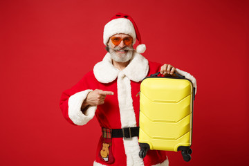 Elderly gray-haired mustache bearded Santa man in Christmas hat sunglasses posing isolated on red background. New Year 2020 celebration concept. Mock up copy space. Pointing index finger on suitcase.