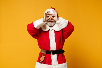 Elderly gray-haired mustache bearded Santa man in Christmas hat isolated on yellow background. New Year 2020 celebration concept. Mock up copy space Hold hand near eye imitating binoculars or glasses.