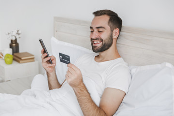 Calm young bearded man lying in bed with white sheet pillow blanket in bedroom at home. Male spend time in room with mobile phone credit card. Rest relax good mood lifestyle concept Mock up copy space