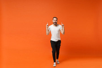 Fototapeta na wymiar Excited young man in casual white t-shirt posing isolated on bright orange wall background studio portrait. People sincere emotions lifestyle concept. Mock up copy space. Pointing index fingers up.