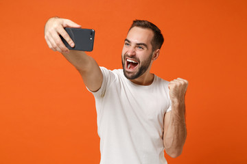 Happy young man in casual white t-shirt posing isolated on orange wall background studio portrait. People lifestyle concept. Mock up copy space. Doing selfie shot on mobile phone doing winner gesture.