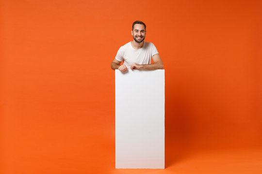 Smiling young man in casual white t-shirt posing isolated on orange wall background. People lifestyle concept. Mock up copy space. Holding big white empty blank billboard with place for text or image.