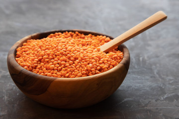 A round wooden bowl filled with red lentils with a wooden spoon stands on a dark background.