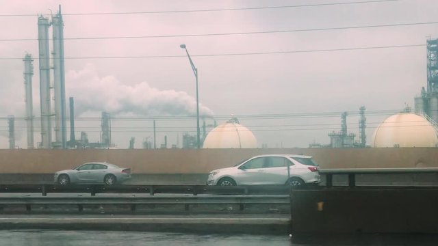 Drving on highway in rain through industrial area with large refineries and factories in background.