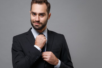 Smiling young business man in classic black suit shirt tie posing isolated on grey wall background studio portrait. Achievement career wealth business concept. Mock up copy space. Straightening tie.