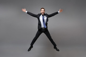 Cheerful young business man in black suit shirt tie posing isolated on grey background. Achievement career wealth business concept. Mock up copy space. Jumping, spreading hands and legs, having fun.