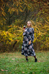 The girl in the dress holds a glass of red wine in the autumn forest