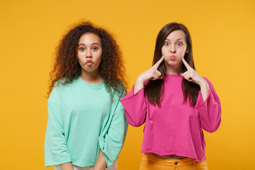 Two women friends european african american girls in pink green clothes posing isolated on yellow background. People lifestyle concept. Mock up copy space. Pointing index fingers on blowing cheeks.