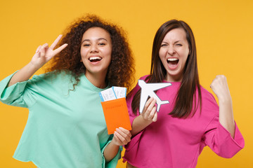 Two women friends european african girls in pink green clothes posing isolated on yellow background. People lifestyle concept. Hold passport ticket airplane showing victory sign doing winner gesture.