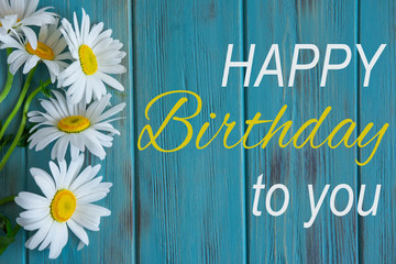 Happy birthday greeting card with camomile flowers on blue wooden background. Flat lay, top view.