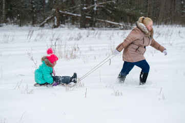 Mother pulling sled with her daughter sitting on it in winter snowy day at nature.