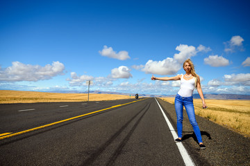 Attractive blond woman with red suitcase hitchhiking on an empty countryside road