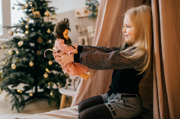 Lovely blonde girl siiting in children room with wigwam and christmas tree and looking at her loving doll in her hands.