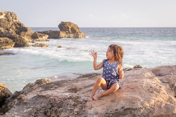 Fototapeta na wymiar Three-year-old girl sitting on a rock in the Caribbean Sea in the morning sun marks three fingers on her hand