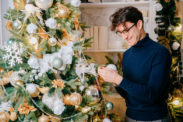 Happy handsome man decorating christmas tree in cozy room with new year holidays decorations.