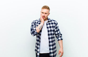 young blonde man with squared shirt holding cheek and suffering painful toothache, feeling ill,...